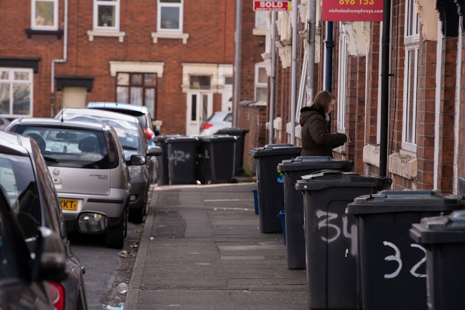 A residential street scene showing a row of black wheeled rubbish bins lined along the pavement outside brick terraced houses with white-framed windows and stone sills. A woman dressed in a dark brown coat is standing next to one of the bins, engaged in handling her waste. Several cars are parked on the street, including a silver hatchback closest to the camera, extending into the background with other vehicles further down the road. The brick buildings feature a mix of traditional design elements, with some signage visible on the walls. Natural daylight illuminates the scene, casting soft shadows on the pavement and highlighting the texture of the brickwork, bins, and parked vehicles. This urban environment illustrates typical domestic waste disposal outside residential properties, which may involve private rubbish collection services, aligning with the context of independent rubbish removal and waste management practices.
