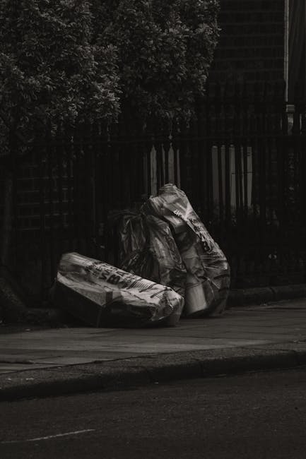 Two large black plastic rubbish bags, filled with waste, are positioned on the pavement near a black metal fence and a wooden gate. One of the bags appears slightly torn at the top, revealing some contents inside. The surface of the bags shows creases and wrinkles, indicating they are tightly packed. Behind the fence, leafy green foliage from a tree is visible, providing a natural backdrop. The scene is set outdoors, in an urban or residential area, with a tarmac road in the foreground. The lighting suggests a cloudy or overcast day, with diffuse natural light casting minimal shadows. This image illustrates typical examples of waste awaiting collection, highlighting the importance of proper rubbish disposal or private waste handling, as services like Rubbish Removal Uxbridge support in managing such waste removal needs.