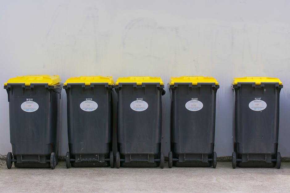 A row of five large wheelie bins made of black plastic with bright yellow lids, positioned against a plain light gray wall in an outdoor setting. Each bin has a white oval label affixed to the front, containing some printed information or barcodes. The bins are aligned on a concrete surface, with the wheels at the bottom visible, and they appear to be empty or awaiting rubbish collection. The environment is well-lit, with natural light evenly illuminating the scene, suggesting daytime. The image conveys a standard waste disposal setup often used for private rubbish collection or on-site clearance, aligning with waste management services like those provided by Rubbish Removal Uxbridge, and demonstrates the type of equipment used for local rubbish storage, ready for collection or disposal as part of alternative waste handling processes.