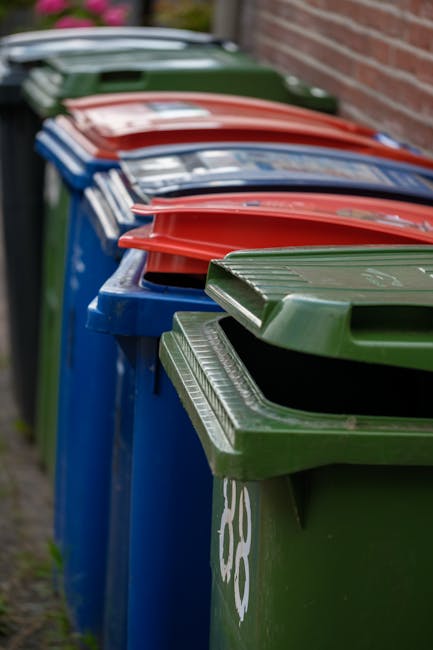 Two large black plastic rubbish bags, filled with waste, are positioned on the pavement near a black metal fence and a wooden gate. One of the bags appears slightly torn at the top, revealing some contents inside. The surface of the bags shows creases and wrinkles, indicating they are tightly packed. Behind the fence, leafy green foliage from a tree is visible, providing a natural backdrop. The scene is set outdoors, in an urban or residential area, with a tarmac road in the foreground. The lighting suggests a cloudy or overcast day, with diffuse natural light casting minimal shadows. This image illustrates typical examples of waste awaiting collection, highlighting the importance of proper rubbish disposal or private waste handling, as services like Rubbish Removal Uxbridge support in managing such waste removal needs.