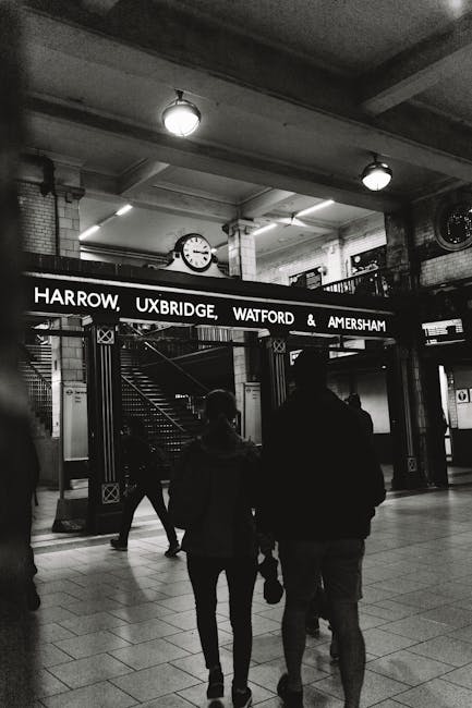 A black and white photograph taken inside a train station shows two individuals walking away from the camera toward an entrance with a sign displaying the destinations Harrow, Uxbridge, Watford, and Amersham. The sign is mounted on a metal frame above the entrance, indicating an underground or train platform. The individuals appear to be carrying bags or personal items, and their silhouettes are visible against the station's illuminated background. The environment includes tiled flooring, brick and metal structural elements, and ceiling-mounted lighting fixtures. In the background, stairs with a metal handrail lead to another level, and a large round clock is mounted on the wall showcasing the time. The scene reflects a typical station setting, where passengers are preparing for or arriving from a journey, and such environments often necessitate efficient rubbish removal and waste management, as handled by companies like Rubbish Removal Uxbridge.