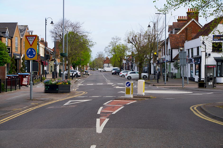 The image depicts a quiet urban street scene with a central pedestrian crossing marked by white dashed lines and red textured pavement. On the left side, a traffic sign indicating a give way junction is visible, alongside a decorative flower bed containing colorful flowers and plants. The street is lined with a variety of shops and cafes housed in buildings with brick and painted facades. Parked cars are positioned along both sides of the street, and street lamps and trees with budding or sparse foliage are visible, suggesting early spring. In the background, more trees and additional parked vehicles define the horizon. The scene is illuminated by natural daylight, with a soft, slightly overcast sky diffusing the light evenly across the area. The photograph appears to capture a typical day for an independent shopping area that may benefit from private rubbish collection or on-site clearance services, supporting the operational needs of local businesses. Rubbish Removal Uxbridge might offer such services to help manage waste efficiently without relying solely on municipal collection.